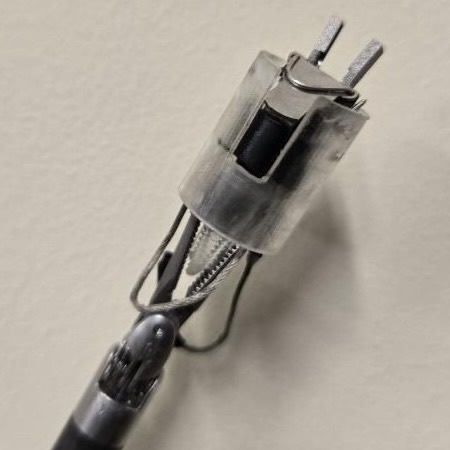 An image of a clip applier prototype being held up by da Vinci Xi Cadiere fenestrated forceps. The prototype is a translucent cylindrical shell with a set of jaws sticking out of the top, and cables hanging loosely from the bottom.