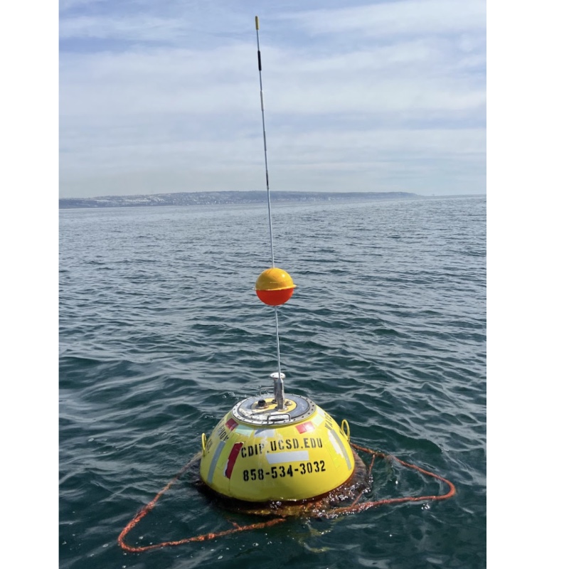 Yellow bouy floating in the ocean with a smaller round orange bouy on a metal rod sticking out the top of the yellow bouy.